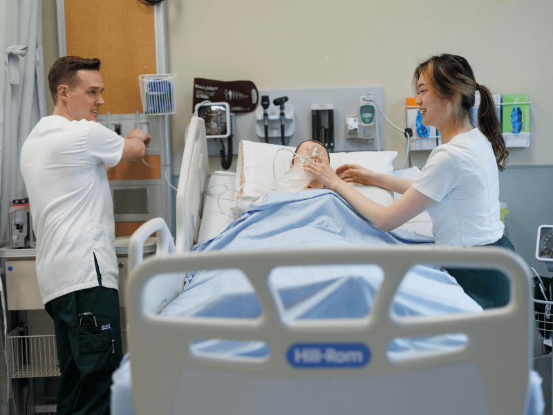 two students caring for a simulated patient in a hospital bed