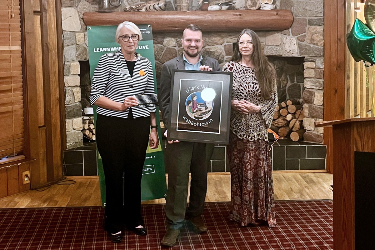 Dean Solina Richter, Kathleen McMullin, and a male are smiling for a photo, holding a piece of art that says USask Nursing wahkohtowin