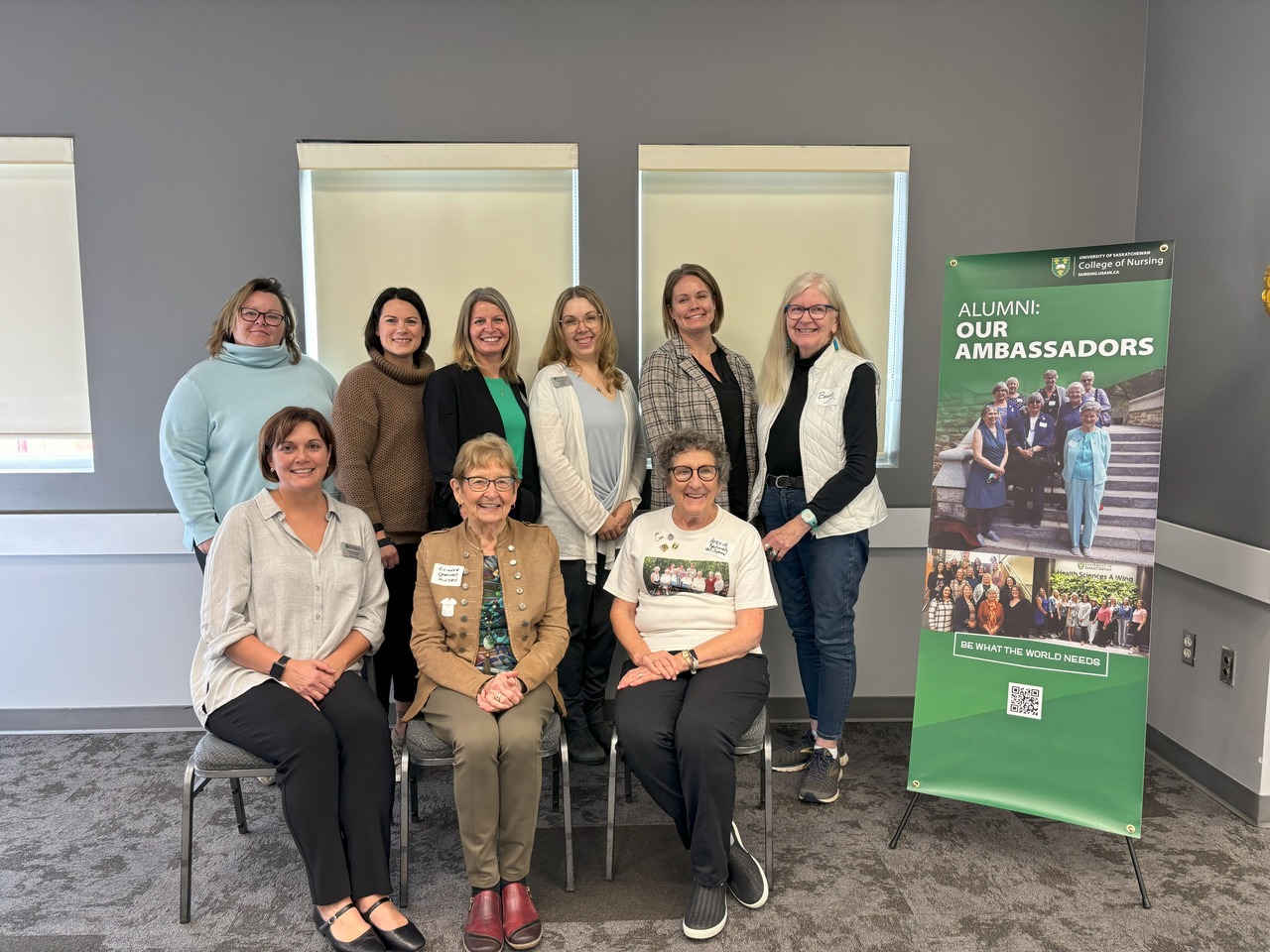 three seated women in front of six standing women, standing beside an Alumni: Our Ambassadors pop-up banner