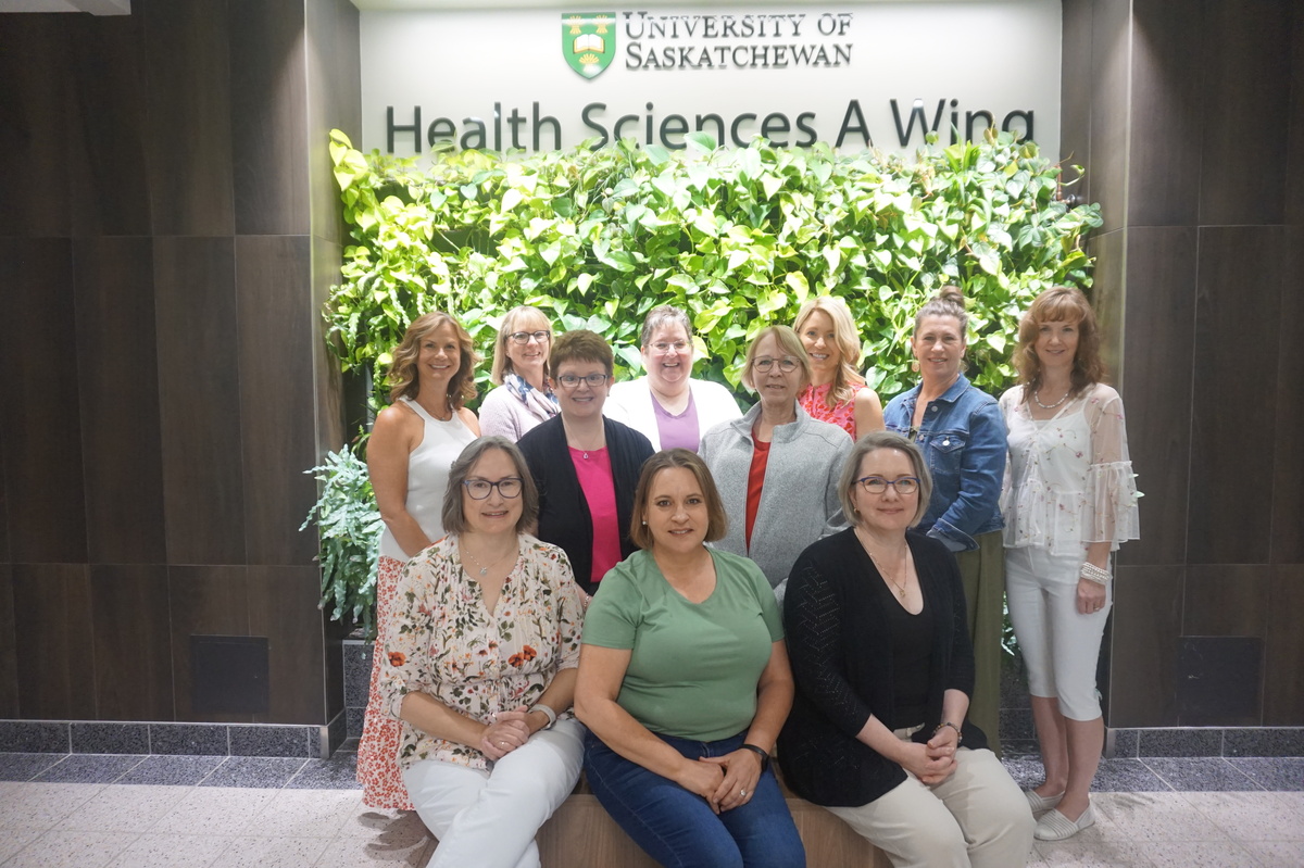 Group photo of the women who attended the 1994 degree class reunion. 3 women sitting on chairs and 8 are standing behind them in front of a wall of plants in the Health Sciences building.