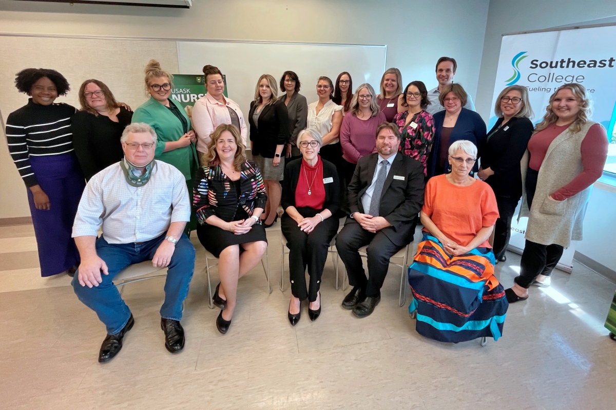 Five people seated in a row smiling. Behind them stands 15 people smiling.