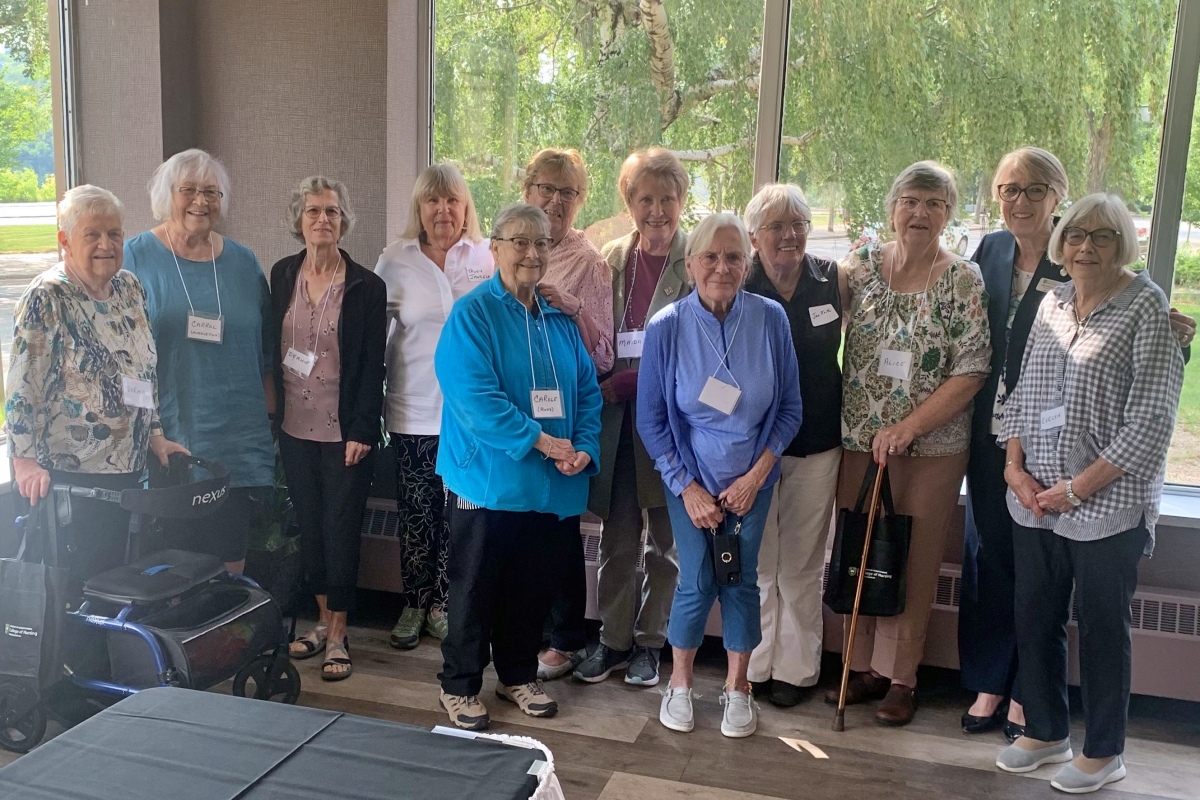 12 women standing together for a group photo, smiling in front of a large window