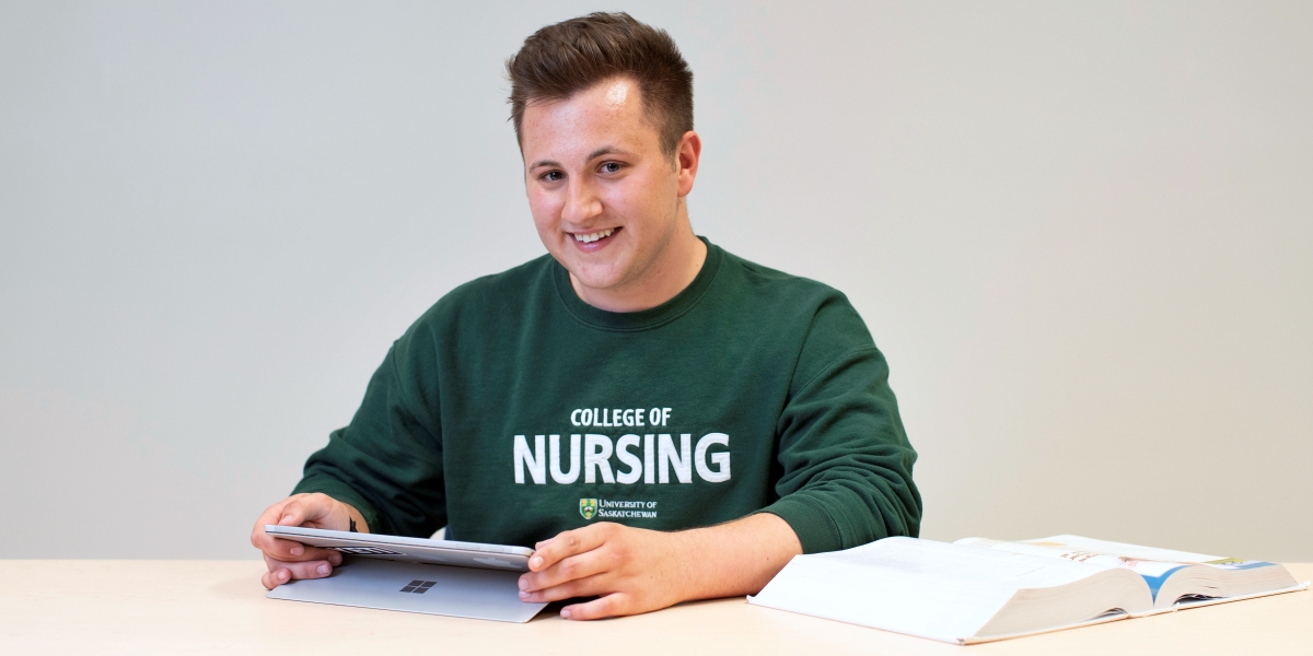 Male student sitting at a table with a textbook, smiling directly at the camera.