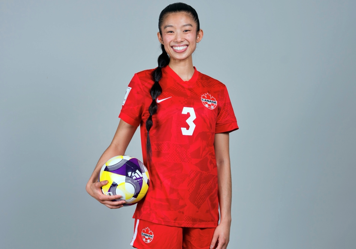 Jade Houmphanh in her Team Canada jersey, holding a soccer ball on her hip and smiling.