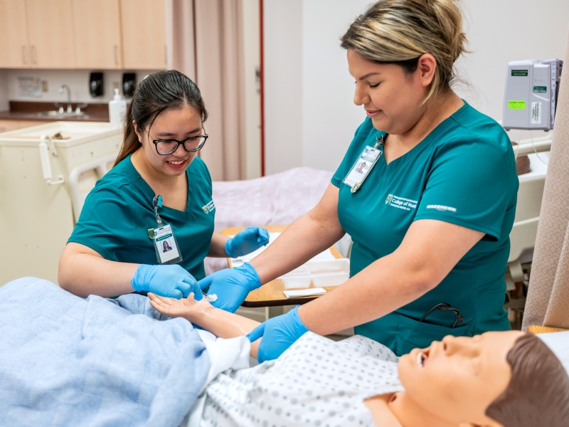 Female students in green nursing scrubs work on mannequin in hospital bed.