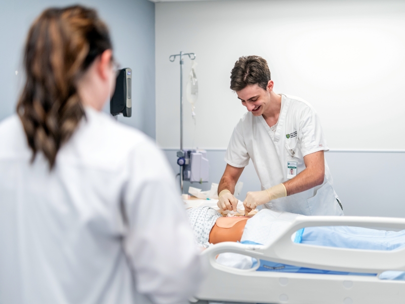 Male student in white nursing scrubs works on mannequin in hospital bed while instructor in white lab coats watches.